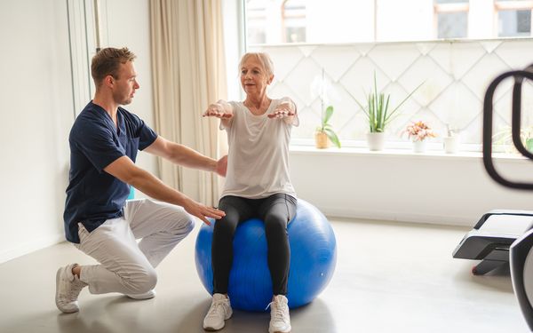 Physiotherapeut zeigt alter Frau Übungen am Gymnastikball