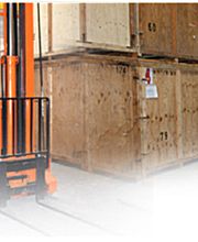 Gabelstapler hebt Holzkiste in Lagerhalle mit Fahrer, umgeben von gestapelten Kisten.