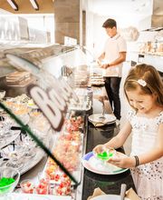 Niña sonriendo mientras elige postres en un buffet, fondo con hombre sirviéndose comida.