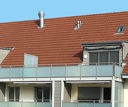 Mehrfamilienhaus mit rotem Satteldach, großen Fenstern und Balkonen, blauer Himmel im Hintergrund.