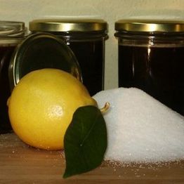 Zitrone und Zucker vor drei dunklen Marmeladengläsern mit goldenen Deckeln auf Holzoberfläche.