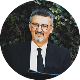 Mann mit Brille und Anzug sitzt vor Efeu, lächelnd, mit offenem Buch in den Händen.