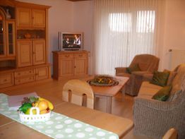 Gemütliches Wohnzimmer mit Holzmöbeln, Sofa, Sessel, Esstisch und eingeschaltetem Fernseher.