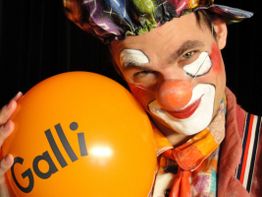 Ein Clown mit weiß geschminktem Gesicht hält einen orangefarbenen Ballon mit "Galli".