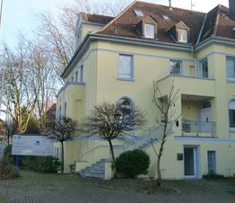 Gelbes zweistöckiges Gebäude mit Treppe und kahlen Bäumen, schattiger Hof mit einer Werbetafel.