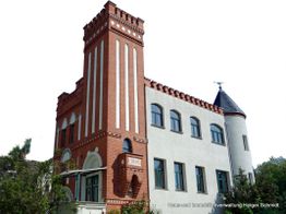 Altes Gebäude mit rotem Backsteinturm und grauer Fassade, umgeben von Bäumen.
