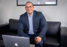 Ein Mann im Blazer sitzt auf einem Sofa mit einem Laptop vor sich und lächelt in die Kamera.