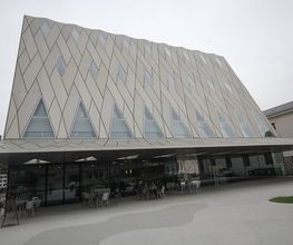 Modernes Gebäude mit schräger Fassade und dreieckigen Fenstern, Menschen sitzen draußen.