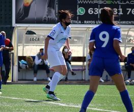 Jugadoras de fútbol en partido, una de blanco y otras de azul, en un campo con banquillo detrás.
