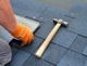 Person installing roof shingles, wearing orange gloves, with a hammer nearby.
