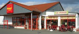 Penny-Markt mit rotem Dach und angrenzendem Imbiss mit roten Außenstühlen und -tischen.