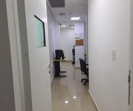 Corredor de escritório com mesas, cadeiras e paredes brancas. Porta aberta em primeiro plano.