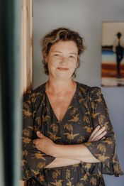 Eine Frau mit lächelndem Gesicht steht vor einer Wand, die Arme vor der Brust verschränkt.