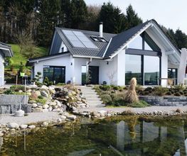 Modernes Einfamilienhaus mit großen Fenstern am Teich, umgeben von Bäumen und Garten.