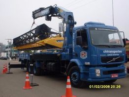 Caminhão azul com guindaste transporta peça grande, cones laranjas ao redor.