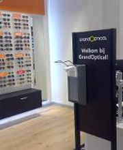 Winkelinterieur met een zonnebrillenwand en balie bij GrandOptical, met een Welkomstbord.