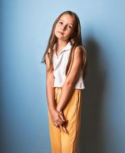 Niña de pie en fondo azul, con camisa blanca y pantalones amarillos, mirando a la cámara.