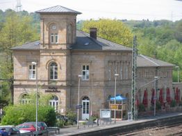 Altes Bahnhofsgebäude aus Sandstein, umgeben von Bäumen, mit Café und Autos im Vordergrund.