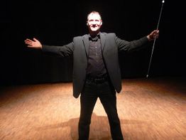 Ein Mann im Anzug auf einer Bühne mit ausgebreiteten Armen, hält einen Zauberstab.