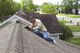 Man repairing shingles on a roof, wearing a hat and jacket, with houses and trees in the background.