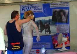 Ein Mann bemalt eine Frau mit Airbrush vor einem Atelier-Weißlein-Werbeplakat.