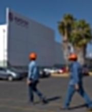 Dos personas con cascos naranjas caminan frente a un edificio blanco y palmeras.
