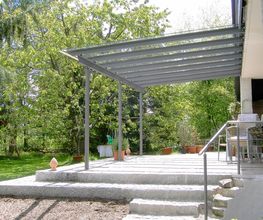 Terrassenüberdachung mit Glasschiebedach im Garten, umgeben von Bäumen und Pflanzen.