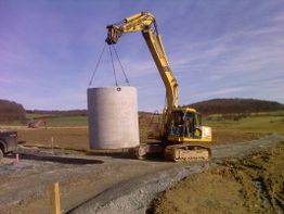 Bagger hebt großen Betonzylinder auf einer Baustelle in ländlicher Umgebung an.
