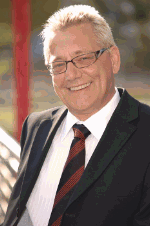 Älterer Mann im Anzug mit Brille, lächelnd, steht draußen bei sonnigem Wetter.