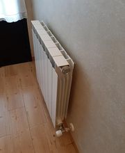 Radiador blanco en una pared de habitación con suelo de madera clara y cortina blanca al fondo.