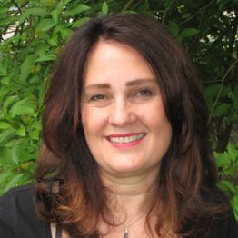 A woman with brown hair smiles in front of green foliage.