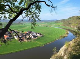 Kleines Dorf an einem Fluss in grüner Landschaft, umgeben von Feldern und Hügeln unter blauem Himmel.