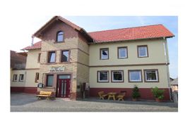 Zweistöckiges Haus mit rotem Dach, gelber Fassade, Holzbänken und Pflanzen vor dem Eingang.