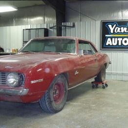 A dusty red classic car in a garage beside a sign that reads "Yankee Automotive."