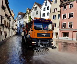 Straßenreinigungsfahrzeug sprüht Wasser in einer Kopfsteinpflasterstraße vor bunten Gebäuden.