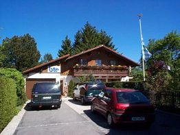 Alpenhaus mit Holzfassade, Autos im Vordergrund, blau-weiße Flagge im Garten, sonniges Wetter.