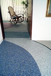 Flur mit blauem und grauem Boden, dahinter ein Raum mit Schaukelstuhl und Pflanze.