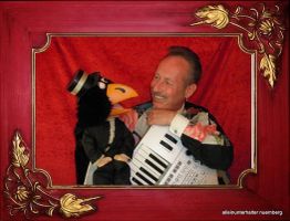 Ein Mann mit Akkordeon lacht mit einer Vogel-Handpuppe vor rotem Hintergrund in Goldrahmen.