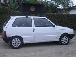 Carro branco com alto-falante grande no teto, estacionado em rua de terra.