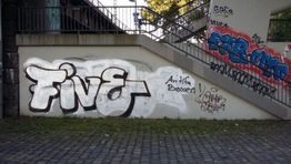 Graffiti unter Treppe: "Five" in Weiß mit Schatten und andere bunte Tags an der Wand.