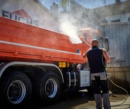 Mann reinigt roten Lkw mit Hochdruckreiniger im Freien neben Gebäude mit Fuchs-Logo.