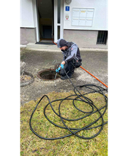 Ein Arbeiter reinigt ein Abflussrohr mit einem Wasserschlauch vor einem Gebäude.