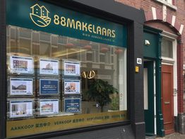Etalage van een makelaarskantoor met woningfoto’s en Chinees en Nederlands opschrift.