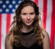 Young woman with long hair smiles slightly, standing in front of a blurred American flag background.