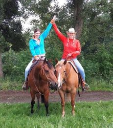 Zwei Frauen auf Pferden klatschen sich ab, umgeben von grüner Natur und Bäumen.