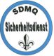 Abzeichen mit blauem Rand, Schriftzug "SDMQ Sicherheitsdienst" und einem Fleur-de-Lis-Symbol.