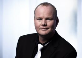Ein Mann im Anzug mit Krawatte vor einem verschwommenen Hintergrund.