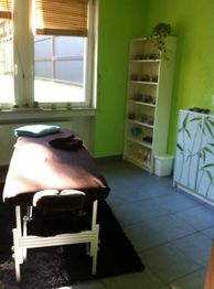 Massageraum mit grün gestrichenen Wänden, Massageliege und Regal voller Handtücher und Produkte.