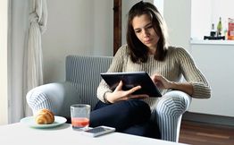 Frau sitzt auf Sessel, nutzt Tablet; auf dem Tisch Croissant und Saft.