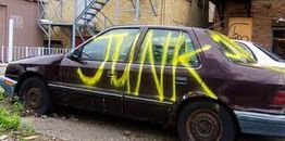 Old car with "JUNK" spray-painted in yellow on the side, parked beside urban buildings.